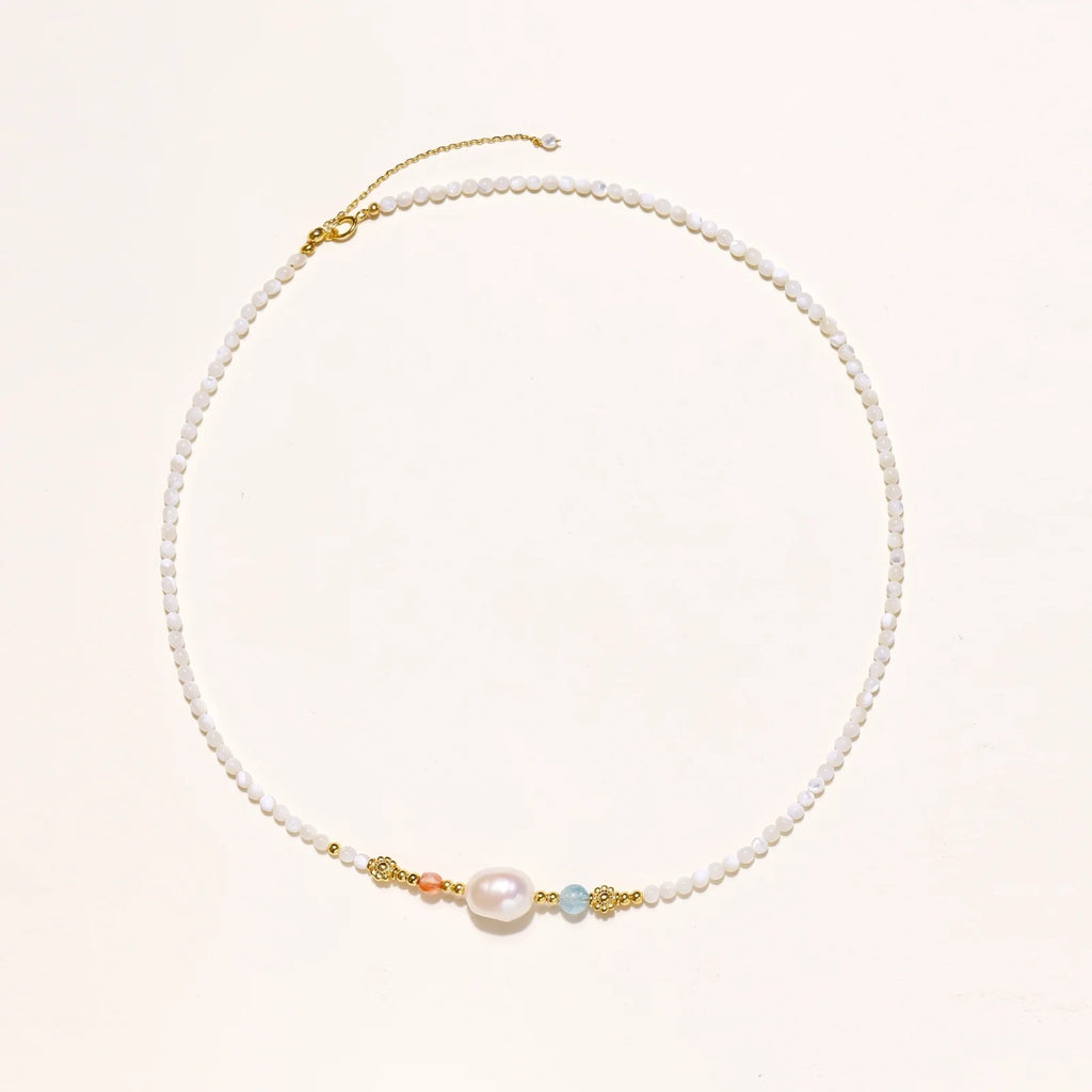 Delicate white beaded necklace with baroque pearl center, gold accents, and coral and aqua gemstones on cream background.