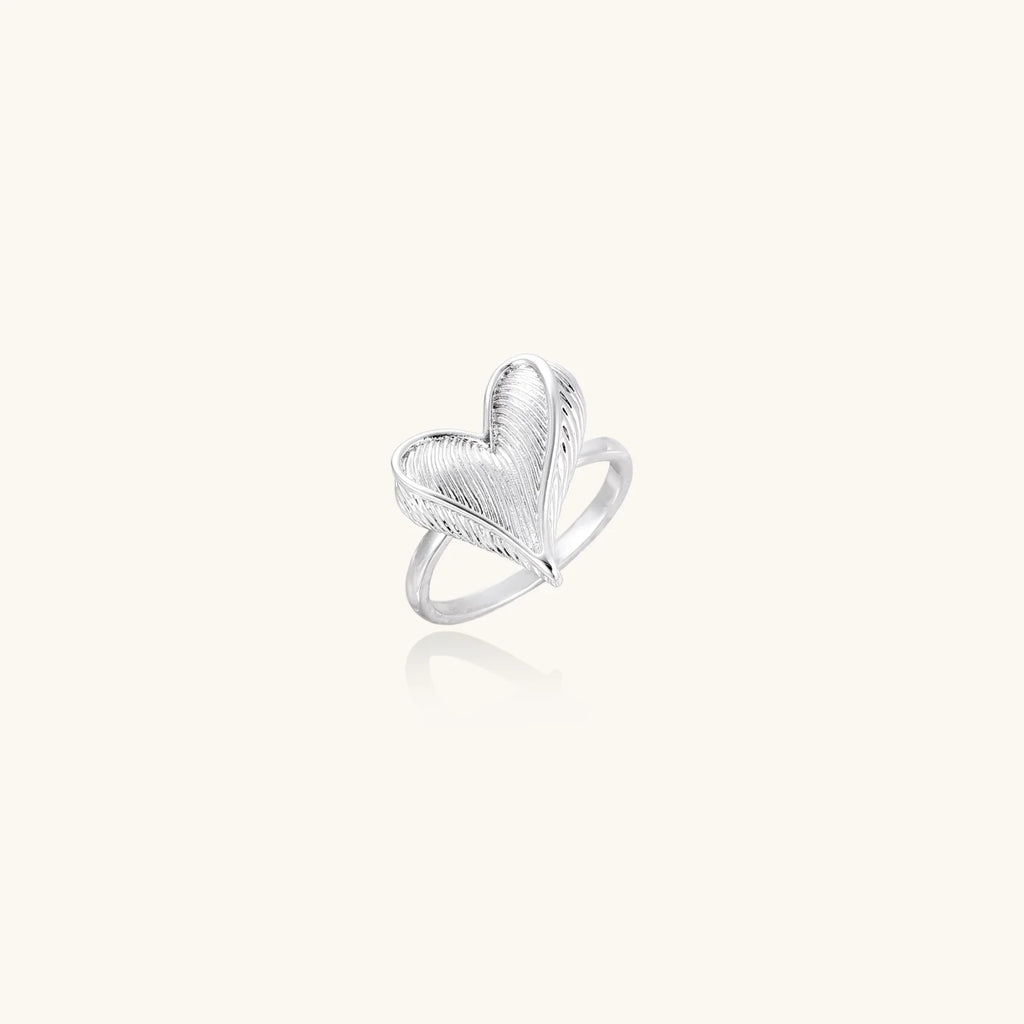 Silver ring with 3D heart formed by textured feather-like elements, white gold plated eco-brass on neutral background.