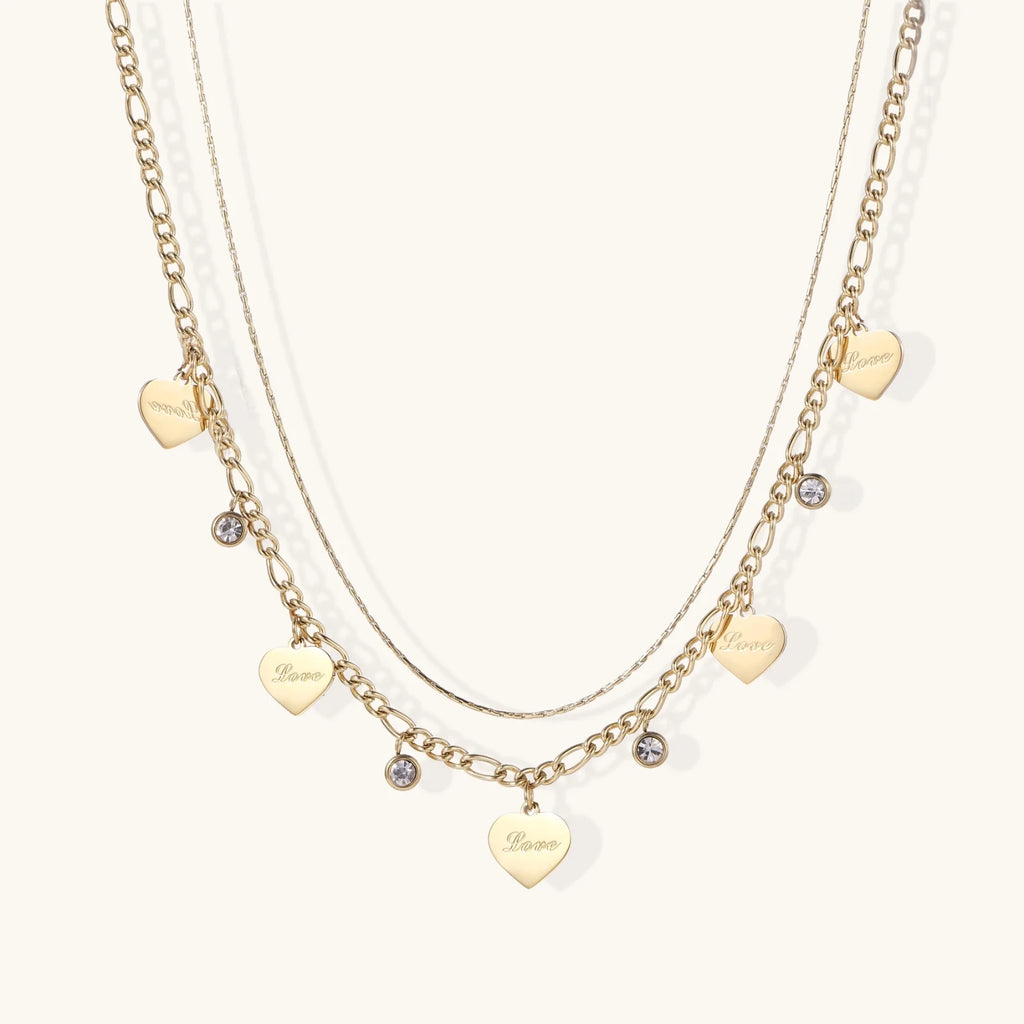 Gold layered necklace set with engraved heart pendants and crystal accents on cream background