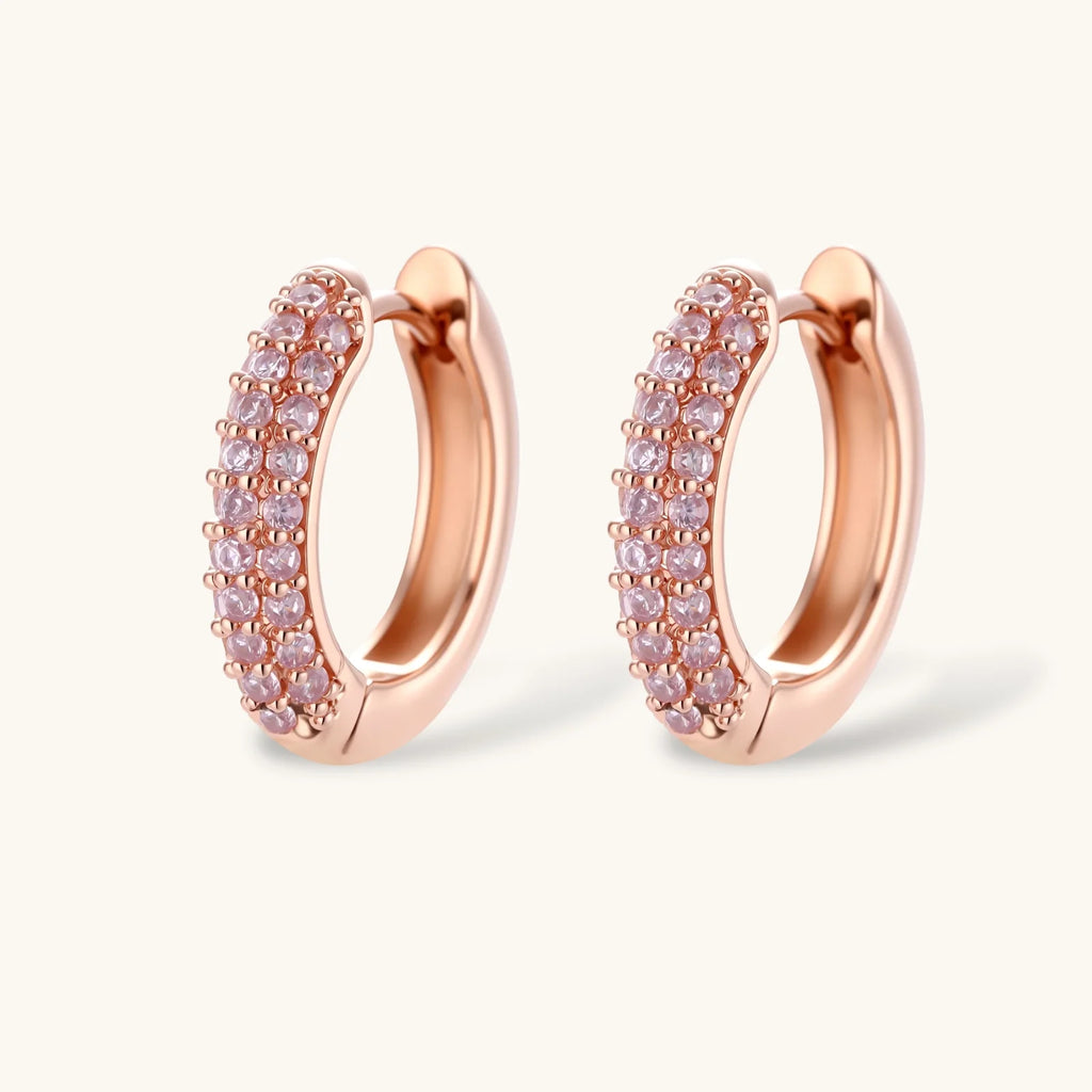 Rose gold jewelry set with pavé cubic zirconia stones including curved necklace, hoop earrings, and adjustable ring on cream background.